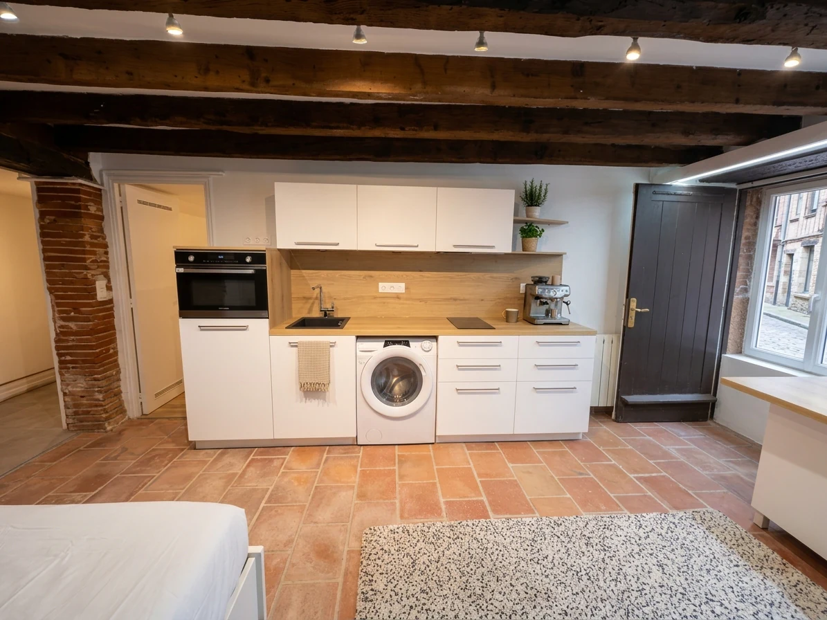 Coin cuisine blanc dans un studio, sous poutres apparentes, mur de briques et sol en tomettes, réalisation MT Cuisine à Toulouse