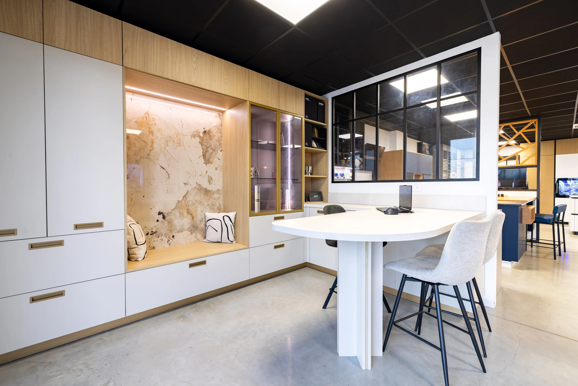 Espace de conception du showroom avec rangements blancs et bois, niche banquette à fond effet pierre, table à bout arrondi et verrière d'atelier