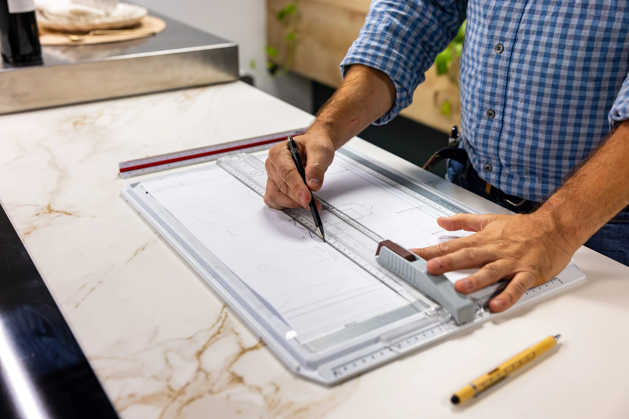 Mains traçant à la règle le plan d'une cuisine sur une planche à dessin posée sur un plan de travail effet marbre
