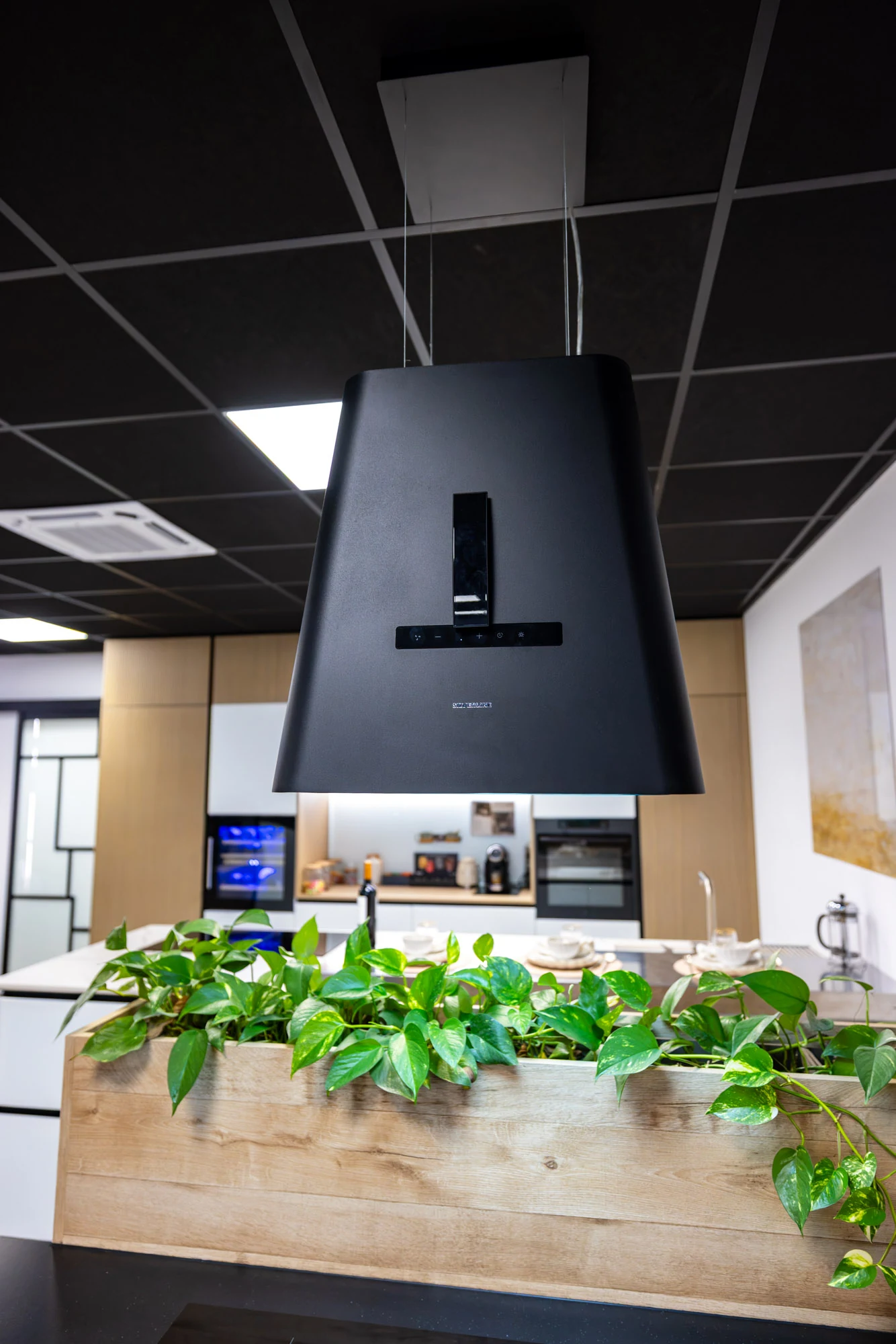 Hotte îlot noire suspendue au-dessus d'une jardinière en bois et de plantes vertes, cuisine blanche et bois en arrière-plan