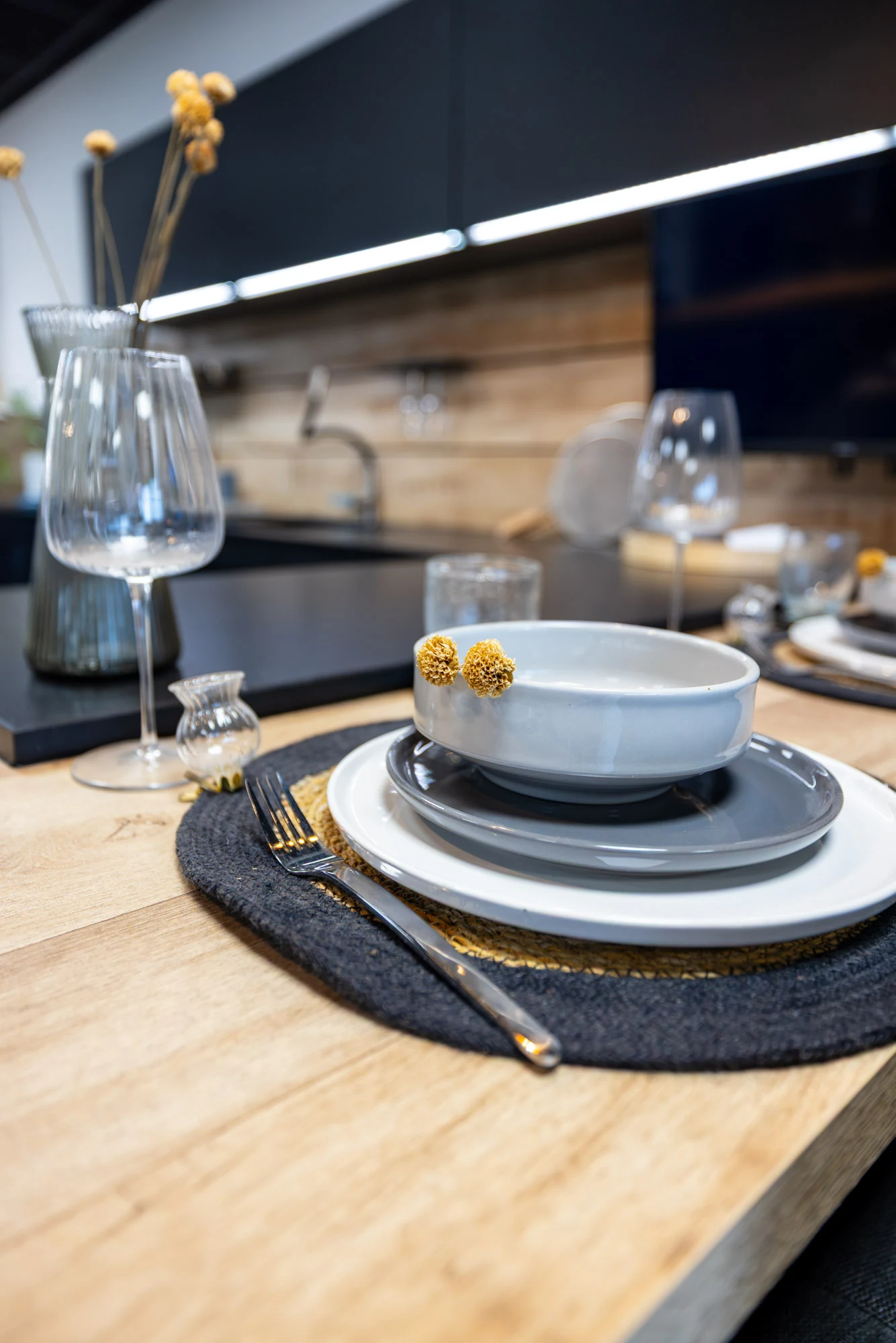 Assiettes grises, verres à pied et set de table tressé sur un plan en bois clair, cuisine noire éclairée en arrière-plan