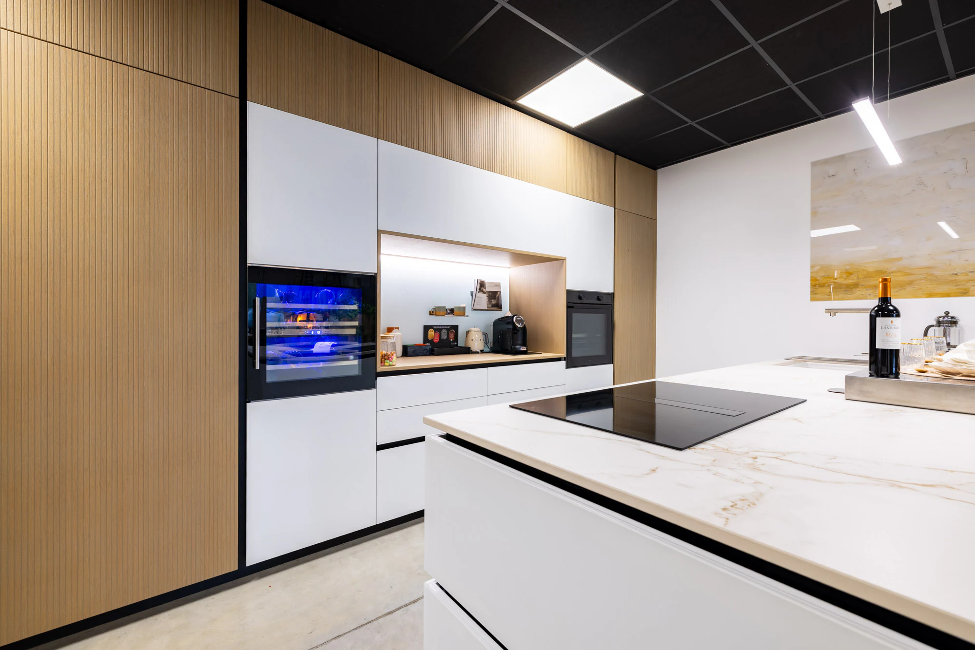 Cuisine blanche à îlot, plan veiné beige et colonnes en bois clair avec four et cave à vin, showroom MT Cuisine à Portet-sur-Garonne