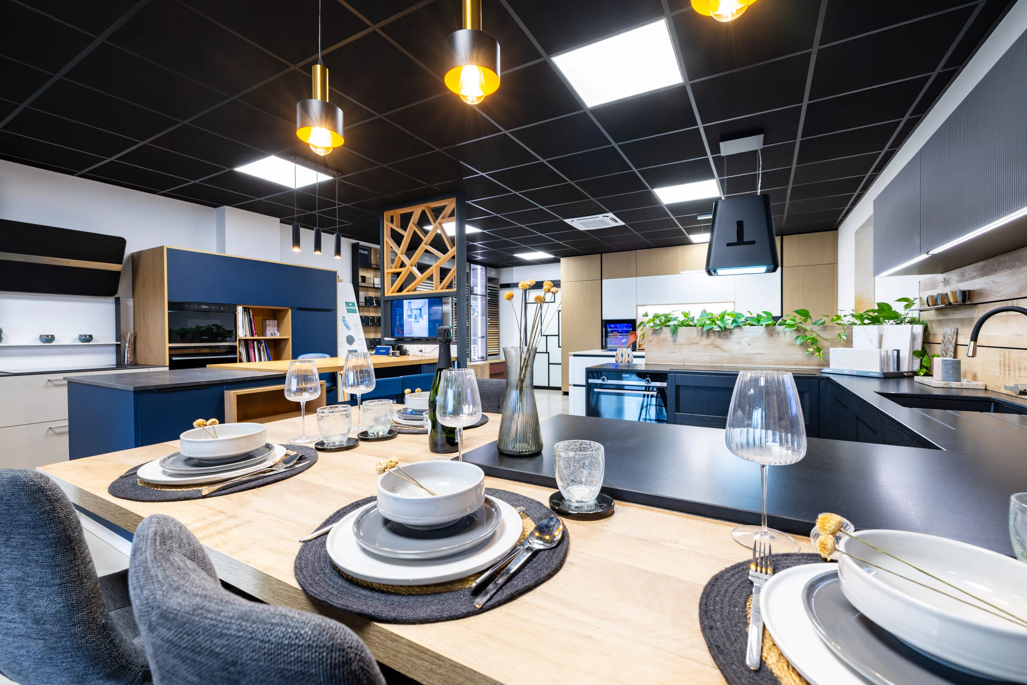 Table en bois dressée au premier plan devant une cuisine bleu marine à îlot et hotte suspendue, showroom MT Cuisine à Portet-sur-Garonne