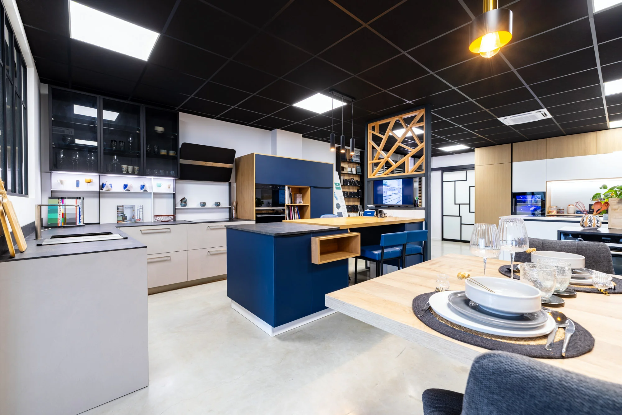 Îlot bleu devant une cuisine grise à vitrines noires et claustra en bois au showroom MT Cuisine de Portet-sur-Garonne