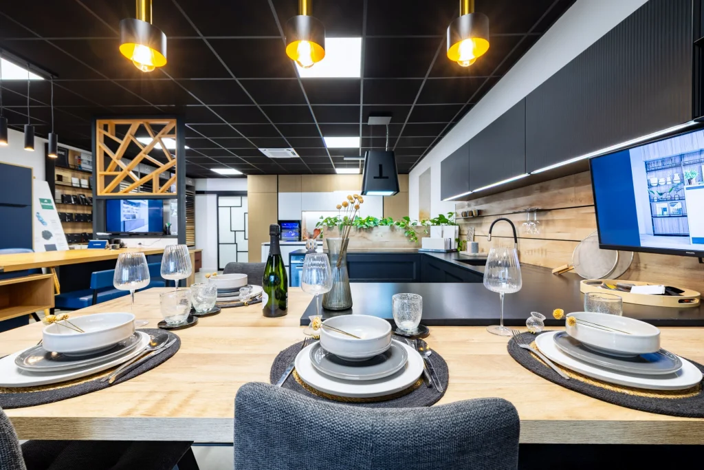 Table haute en bois dressée devant une cuisine noire, suspensions laiton et écran de présentation au showroom MT Cuisine de Portet-sur-Garonne