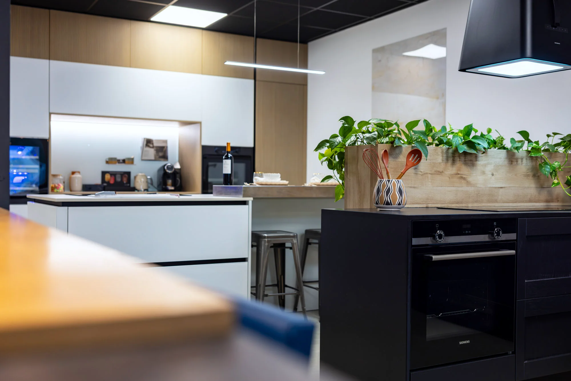 Cuisine blanche à îlot vue depuis un plan en bois, four noir encastré au premier plan au showroom MT Cuisine de Portet-sur-Garonne