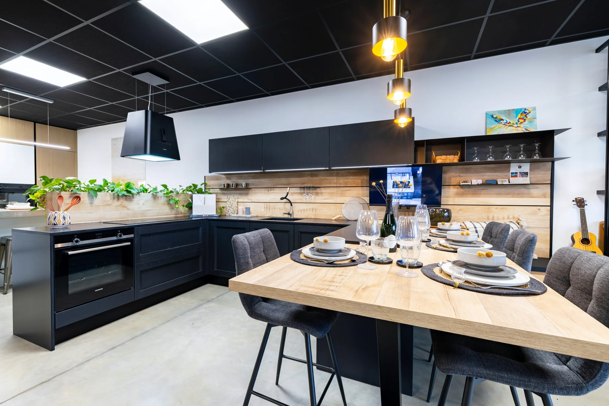 Vue d'ensemble d'une cuisine noire en L avec crédence en lames de bois et table haute en bois dressée au showroom MT Cuisine de Portet-sur-Garonne